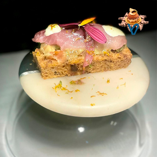 Es un pincho sencillo y muy fresco. Sobre una base de pan de tomate, añadimos una crema de guacamole y sobre ella un trozo de lomo de txitxarro, ahumado por nosotros a los cítricos. Sobre este dos puntos de ali.oli uno de vermú y otro de lima. Con un punto de mermelada de arándano azul.