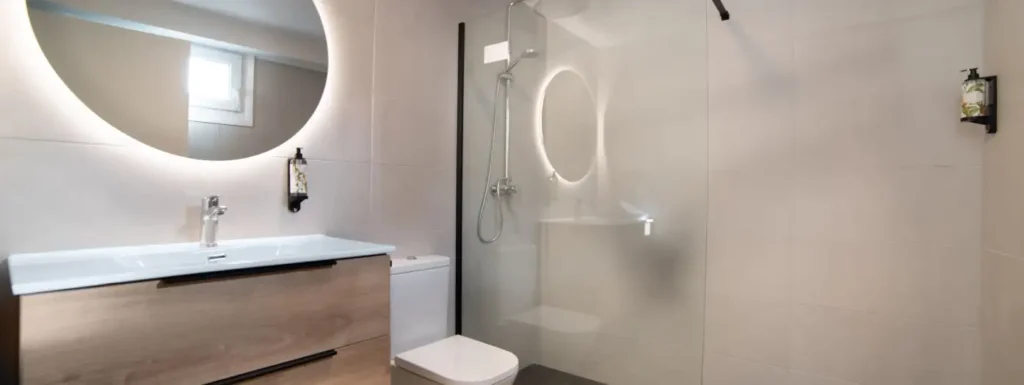 Aseo completo, con ducha y mampara de cristal, aseo y mueble de baño moderno de madera con tiradores en negro. Espejo circular iluminado desde atrás