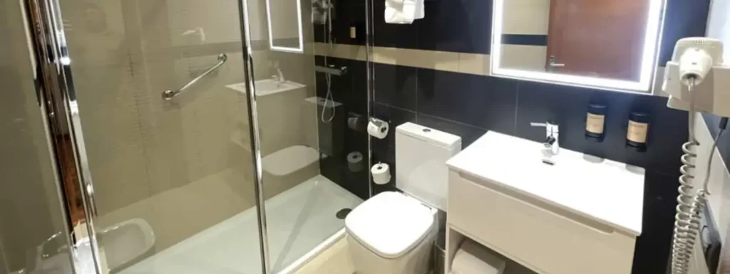 aseo completo del hotel, laba manos y aseo con un gran espejo en la parte derecha y una ducha con mampara transparente al fondo