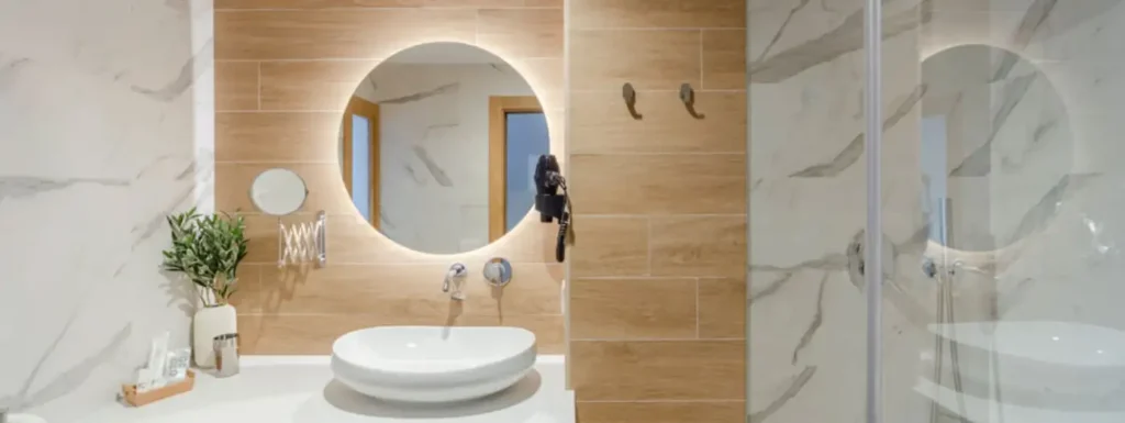 Baño moderno, con marmol blanco y madera en las paredes a la izquierda, el lavabo en altura, con espejo redondo con luz trasera y a la derecha la ducha, con mampara de cristal