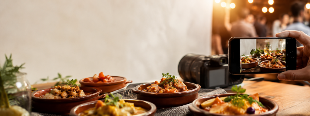 Imagen concurso de fotografía, se ve en una mesa 5 cazuelizas y una camara de fotos, a la vez que una mano soteniendo un movil está enfocando las cazuelicas para hacer una fotografía.