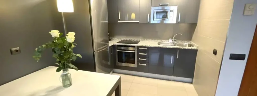 Cocina completa, dos rosas blancas, en una mesa, en primer plano en el fondo de izquierda a derecha, nevera, horno-vitro, cajoneras, y lavabo. Sobre la encimera campana y micro hondas