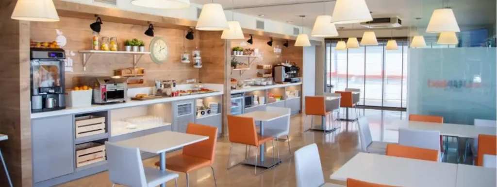 espacio del desayuno buffet. Mesas con sillas naranjas distribuidas por el salón, en el fondo barra desayunador