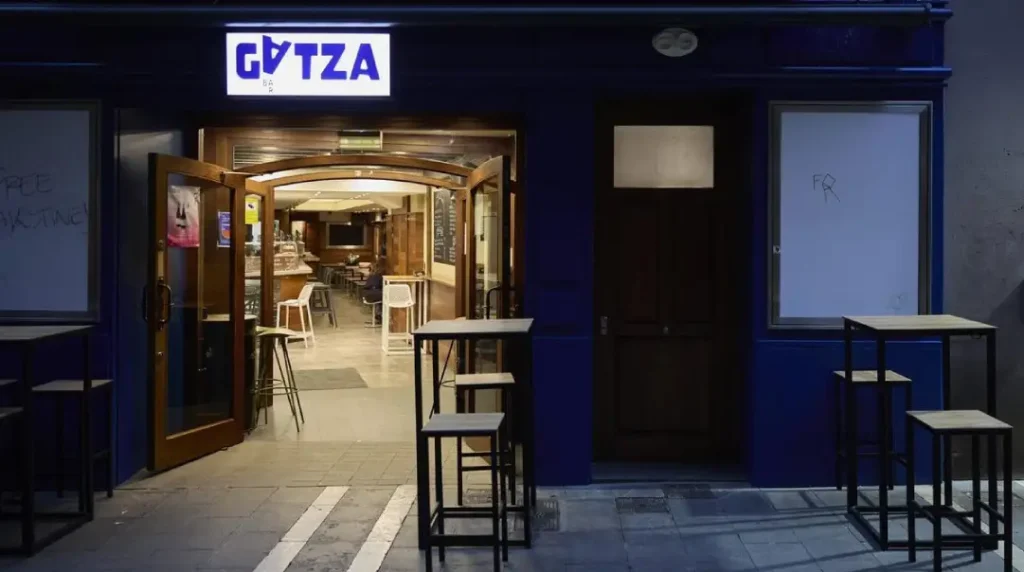 Entrada del bar Gatza, paredes en azul, con dos mesas altas a los lados de las puertas, con sillas en formato taburete alto. El letrero del bar en luminoso con azul