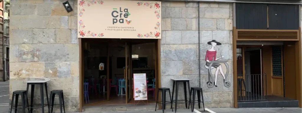 cartel floral en tonos pastel con el nombre del establecimiento, porton de madera con gran cristalera. Mesas alatas con taburetes a los laterales de las dos puertas.  y un dibujo sobre la piedra de una mujer tomando un cafe