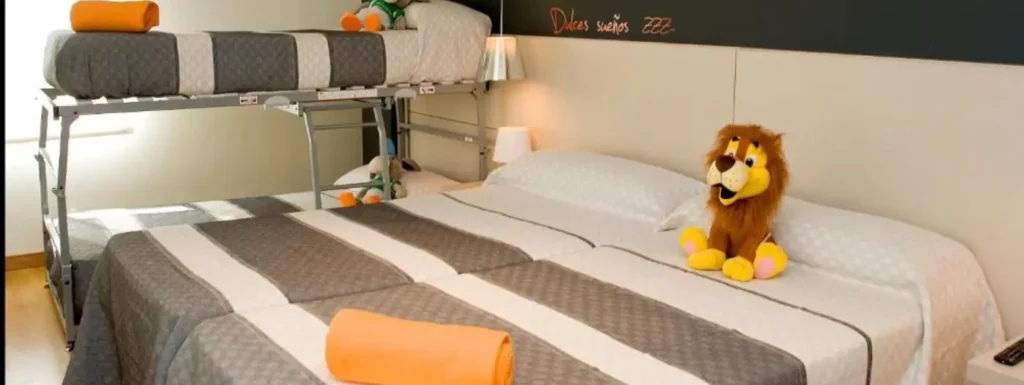 Habitación con dos camas individuales y literas en el fondo. Colcha gris y blanca a rayas, sobre la cama un peluche de un león