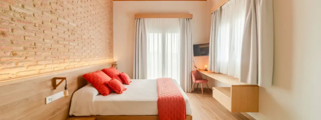 habitación de aspecto calido, parez de la izquierda de piedra, con iluminación en led, desde el cabecero d la cama, que es doble con detalles en naranja y colcha blanca. Frente la cama un escritorio