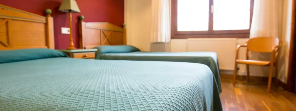 habitación doble, con dos camas con colchas verdes. Pared del cabecero pintada de rojo y cabeceros de madera. al fondo una ventana con una silla de madera