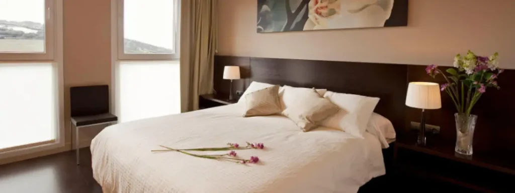 habitación de cama grande, con dos ventanas hasta el suelo al fondo y un cabecero en madera oscura, con colcha blanca cojines y dos flores decorativas sobre la colcha