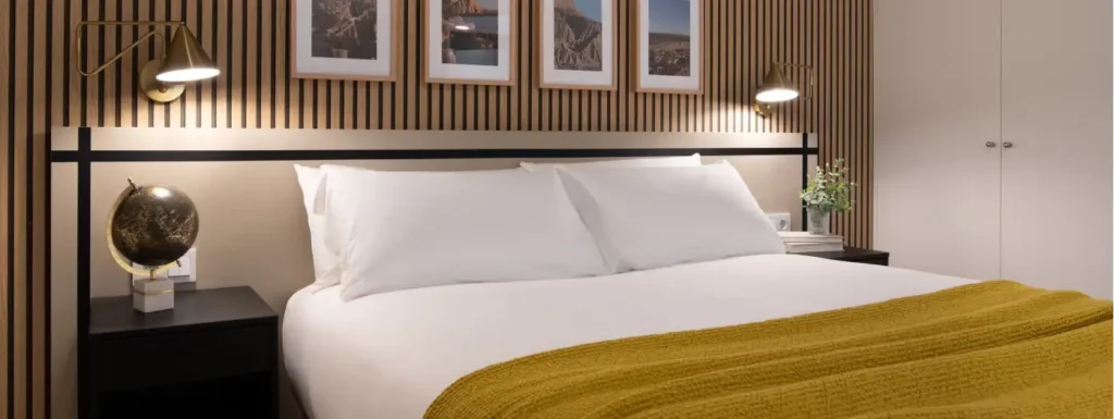 Cama de matrimonio con colcha amarillo mostaza, pared laminada de madera con cuatro cuadros sobre el cabecero de la cama