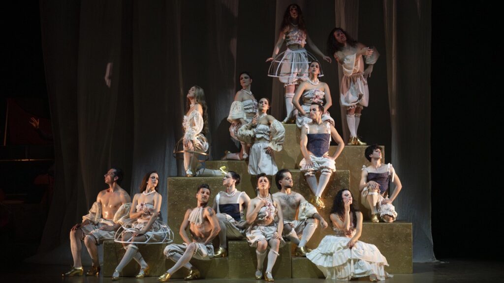 Imagen escénica de un espectáculo teatral o musical con un grupo de intérpretes distribuidos sobre una estructura de gradas doradas. Visten trajes de estética barroca o cabaretera, en tonos claros, con corsés, volúmenes, perlas, medias y zapatos dorados. La composición crea una imagen coral muy visual, con poses marcadas y expresivas, iluminación cálida y un fondo oscuro de telones que refuerza el carácter teatral. El conjunto transmite elegancia, exceso escénico, sensualidad y una puesta en escena cuidada, con una clara intención estética y dramática.