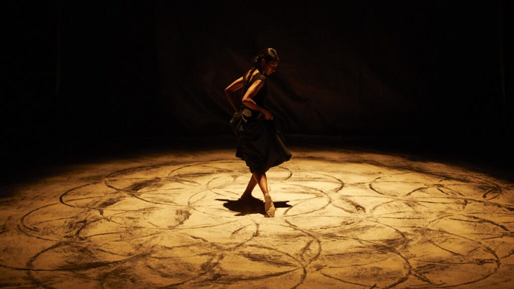Bailarina en un escenario oscuro sobre una zona circular iluminada permanece en pie,