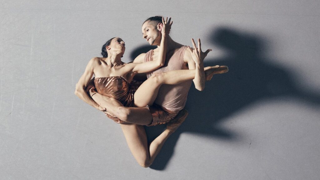 Imagen de danza contemporánea con dos intérpretes en una composición corporal muy precisa y escultórica. La bailarina aparece sostenida en brazos del bailarín, con una pierna elevada y el cuerpo girado hacia él, generando una sensación de tensión, equilibrio y confianza. Ambos visten prendas en tonos cálidos y satinados, que contrastan con el fondo gris neutro. La iluminación lateral proyecta sombras marcadas y refuerza la expresividad física de la escena. El conjunto transmite intimidad, fuerza, delicadeza y una clara conexión entre movimiento, técnica y emoción.