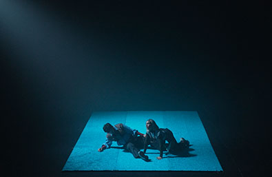 Escena escénica en un espacio oscuro, con dos intérpretes sobre una plataforma iluminada en tonos azules. La composición crea una atmósfera íntima y experimental, con el resto del escenario en penumbra.