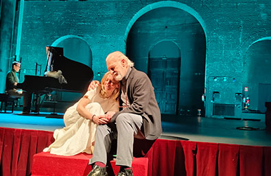 Escena teatral con dos intérpretes sentados en una tarima roja, acompañados por un pianista al fondo, en un escenario iluminado en tonos azulados.