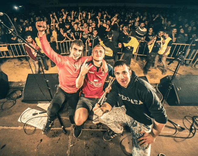 Tres integrantes de la banda Dinamita posan sobre el escenario frente al público tras un concierto, rodeados de micrófonos, cables y equipos de sonido.