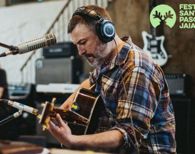 Músico tocando una guitarra acústica con auriculares, sentado junto a varios micrófonos en un escenario. Viste camisa de cuadros y aparece concentrado durante la actuación, con equipos de sonido y un logotipo de festival al fondo.