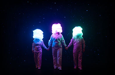 Tres intérpretes de pie y cogidos de la mano en un escenario oscuro con fondo estrellado. Sus cabezas aparecen cubiertas por volúmenes luminosos de colores azul, rosa y verde, creando una imagen escénica de carácter fantástico y visual.