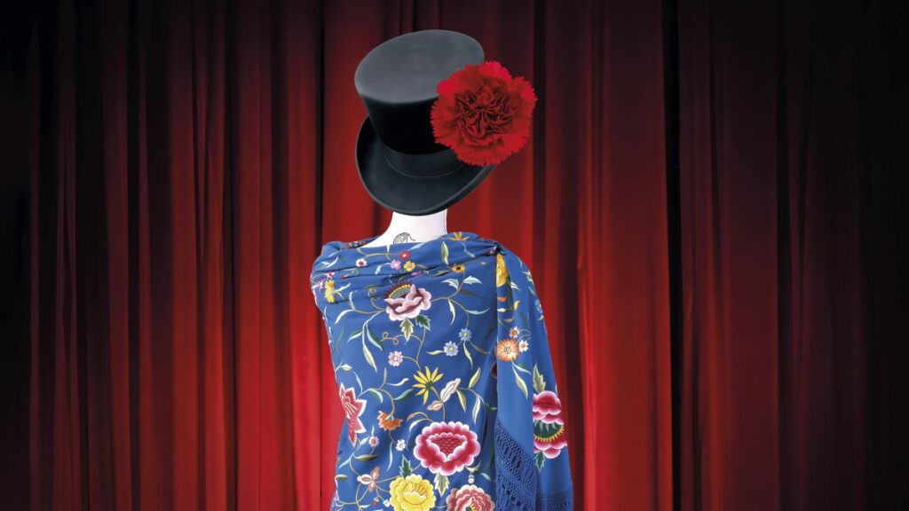 magen de estética teatral y folclórica, centrada en una figura sin rostro vestida con un mantón azul intenso bordado con flores de colores y largos flecos. Sobre la parte superior aparece un sombrero negro con un clavel rojo, reforzando la referencia a la tradición escénica y popular. El fondo de cortinas rojas, los claveles en el suelo y la sombrilla blanca apoyada junto a la figura crean una composición elegante, simbólica y muy visual. El conjunto transmite teatralidad, identidad cultural, color y una fuerte presencia escénica.