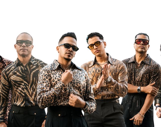 rupo de cinco hombres posando de frente sobre fondo blanco, con gafas de sol, camisas estampadas de estilo animal print y pantalones oscuros. La imagen tiene un carácter promocional y muestra una estética urbana y coordinada.