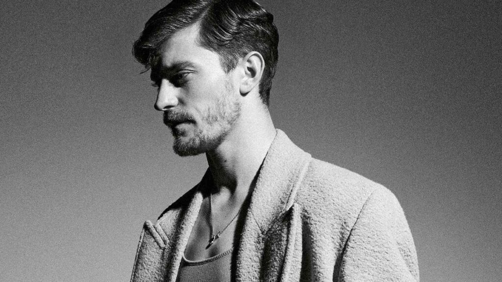 Retrato en blanco y negro de un hombre joven en plano medio, fotografiado de perfil y con la mirada dirigida hacia abajo. Viste una chaqueta de textura gruesa sobre una camiseta de punto, con una estética sobria, elegante y contemporánea. La iluminación lateral marca el rostro y aporta profundidad, mientras el fondo neutro refuerza el carácter íntimo y editorial de la imagen. El conjunto transmite serenidad, introspección y una imagen cuidada, cercana al retrato artístico o promocional.