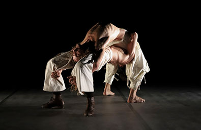Dos bailarines en una escena de danza contemporánea, entrelazados en una postura acrobática sobre un escenario oscuro.