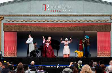 Representación escénica al aire libre sobre un escenario móvil del Teatro Real, con varios intérpretes caracterizados con vestuario de época ante el público. En la parte superior del escenario aparece el logotipo “Teatro Real, cerca de ti”.