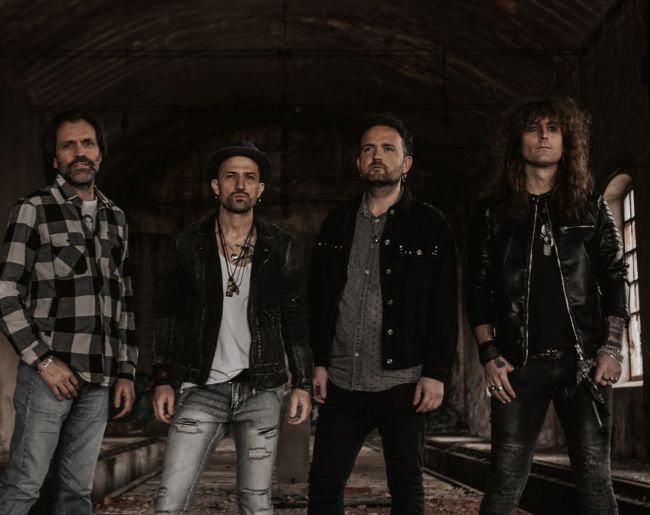 Grupo de cuatro hombres posando de pie en un espacio oscuro con aspecto industrial o abandonado. Visten ropa de estilo rock, con chaquetas de cuero, camisa de cuadros, vaqueros y tonos oscuros, mirando de frente a cámara con actitud seria.