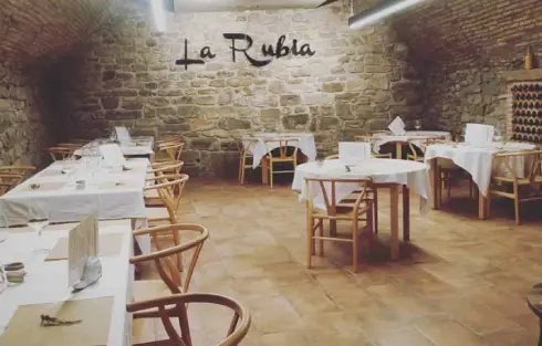 Comedor de piedra con suelo de madera y mesas montadas para el servicio. Cartel de La Rubia en la pared