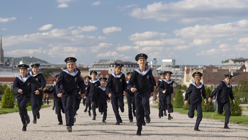 La imagen muestra a un grupo de niños vestidos con uniforme marinero oscuro, corriendo por un camino de grava en un espacio abierto. Van con gorra, cuello marinero blanco y pantalón azul marino. La escena tiene un aire dinámico y alegre, con varios niños avanzando hacia la cámara.