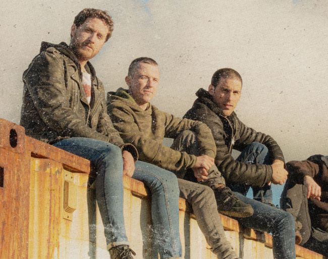 Fotografía de tres hombres sentados sobre una estructura de madera en exterior, con ropa informal y mirada hacia la cámara, en una imagen de estética sobria y urbana.