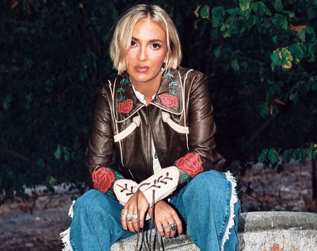Mujer joven rubia sentada en exterior, mirando directamente a cámara. Viste chaqueta marrón con bordados florales rojos y verdes, pantalones vaqueros claros y varios anillos, con vegetación oscura al fondo.