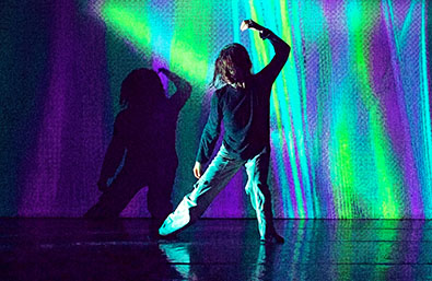 Intérprete en escena realizando una coreografía frente a una proyección de luces verdes, azules y violetas. La figura aparece de espaldas y genera una sombra sobre el fondo, en una imagen de carácter contemporáneo y visualmente envolvente.
