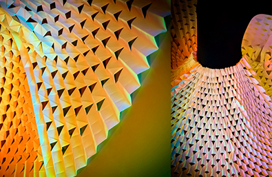 Composición visual con estructuras textiles o escultóricas de formas geométricas repetidas, iluminadas con tonos amarillos, naranjas, verdes y azules. La imagen destaca por su textura, volumen y efecto lumínico, creando una atmósfera abstracta y envolvente.