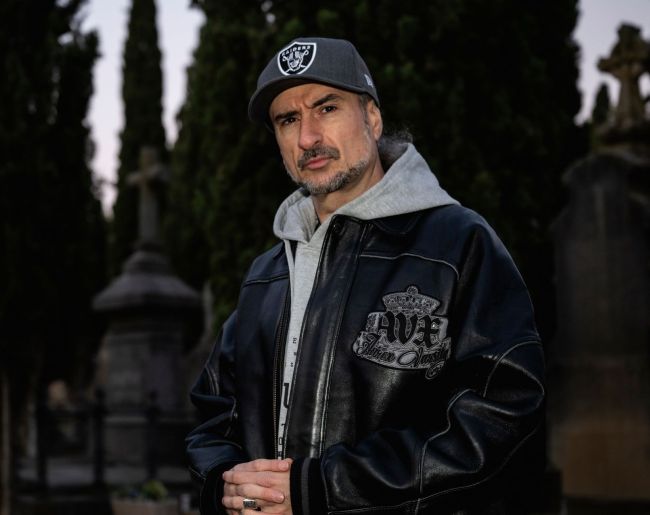 Hombre posando en exterior con gorra gris, sudadera con capucha y chaqueta negra de cuero. La imagen tiene un fondo oscuro y desenfocado con árboles y elementos arquitectónicos, y muestra al protagonista mirando directamente a cámara.