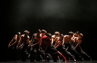 Grupo de bailarines sobre un escenario oscuro, realizando una coreografía contemporánea en formación compacta. La escena está iluminada desde arriba y combina vestuario negro con detalles rojos, creando una imagen de gran intensidad y movimiento.