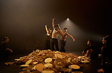 Escena de danza contemporánea sobre un escenario oscuro, con varios intérpretes alrededor de un gran conjunto de panderos y percusiones colocados en el suelo. En el centro, tres bailarines realizan una coreografía bajo una iluminación cálida y focalizada.