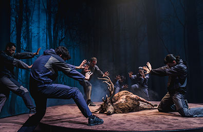 Escena teatral o de danza en un bosque oscuro, con varios intérpretes alrededor de la figura de un animal tumbado en el suelo. Los artistas realizan gestos tensos y expresivos bajo una iluminación azulada, creando una atmósfera dramática.