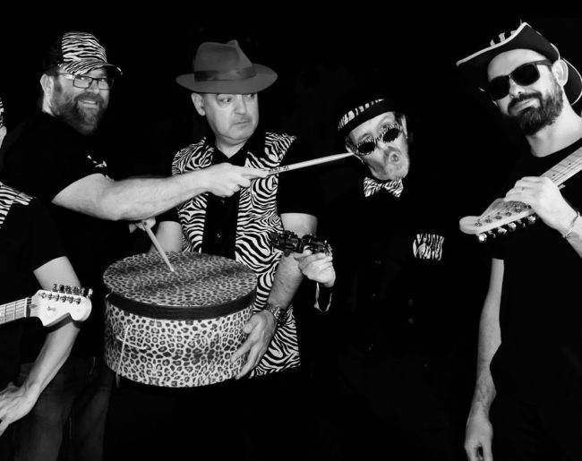 Cuatro músicos posan en blanco y negro con guitarras, percusión y estética desenfadada, vestidos con sombreros, gafas y estampados llamativos.