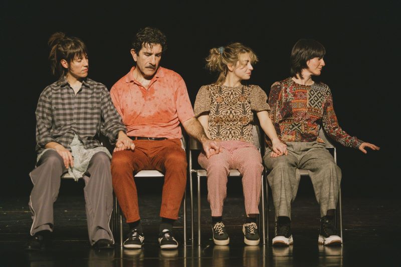 Cuatro intérpretes sentados en fila sobre un escenario durante una representación teatral, vestidos con ropa de estilo informal y tomados de la mano, sobre un fondo negro.