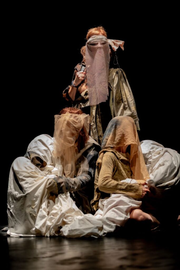 Varias intérpretes sobre un escenario durante una representación teatral, agrupadas y cubiertas parcialmente con telas y velos en tonos blancos y beige, sobre un fondo negro.