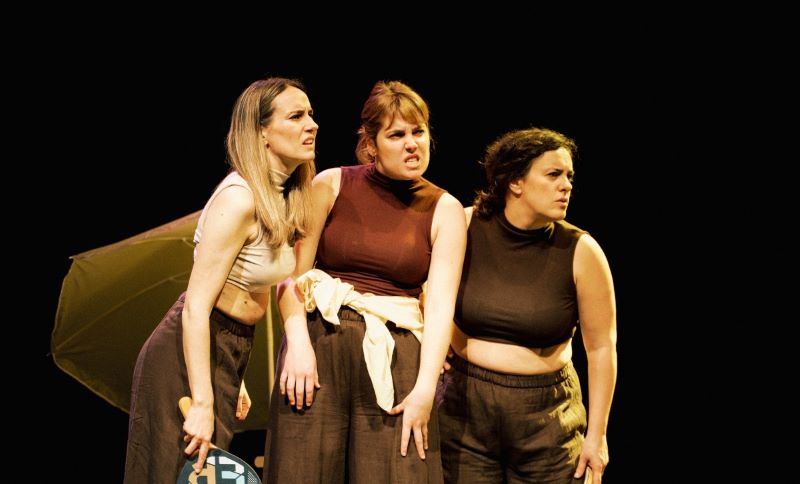Tres actrices sobre un escenario durante una representación teatral, situadas juntas y mirando hacia un mismo punto con expresión de tensión o sorpresa. Al fondo se aprecia parte de la escenografía sobre un fondo negro.