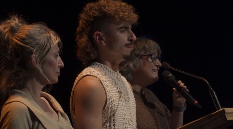Tres personas sobre un escenario durante un acto público. En primer plano aparecen una mujer y un hombre, mientras otra mujer interviene ante un micrófono desde un atril.
