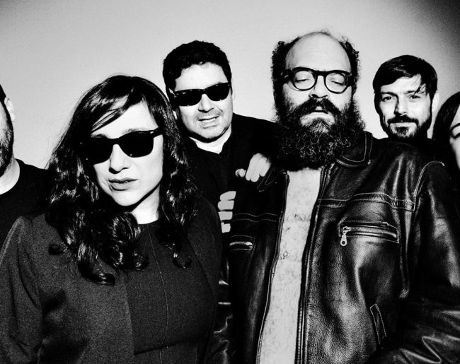 Fotografía en blanco y negro de un grupo musical posando ante la cámara, con varios integrantes en primer plano, algunos con gafas de sol.