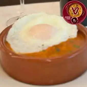 Cazuelica de barro con una elaboración de color anaranjado, cubierta con un huevo frito y decorada con pequeñas hierbas verdes.