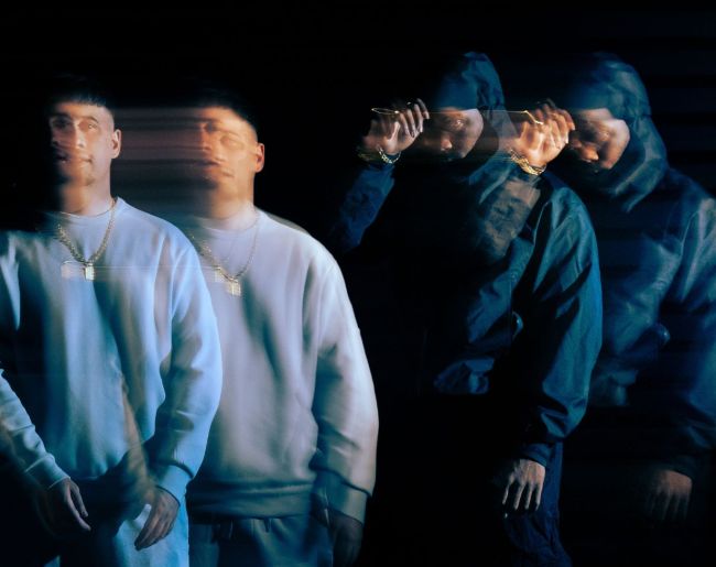 Dos hombres posan sobre un fondo negro con un efecto visual de movimiento y exposición múltiple. Uno viste una sudadera clara y el otro ropa oscura con capucha.