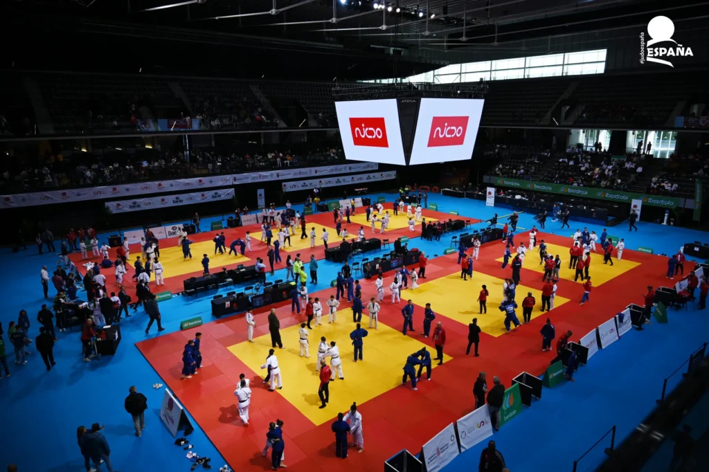 Vista general de una competición de judo en un pabellón deportivo, con varios tatamis ocupados por judocas durante los combates y numeroso público en las gradas.